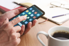 Person using a smartphone with app icons visible while working at desk with notebook and coffee cup in view.