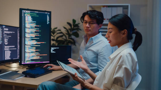 Developers discussing mobile app code with tablet and multiple monitors in a home office environment.